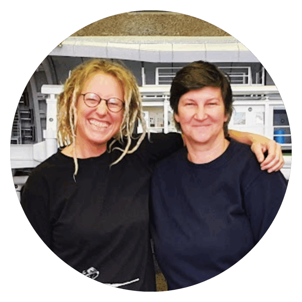 Gesa Witt und Tine Lippert schauen gemeinsam in die Kamera. Sie stehen vor einem technischen Objekt, das sie restaurieren.