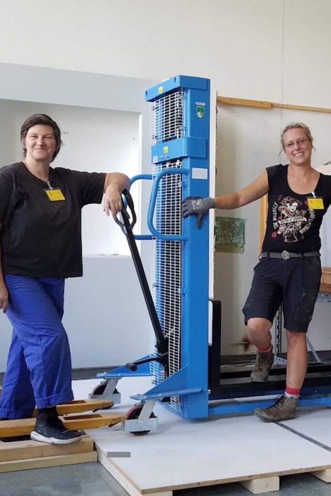 Tine Lippert und Gesa Witt mitten bei der Arbeit mit einem Hubwagen. Sie schauen in die Kamera. Sie sind der richtige Kontakt für Restauratoren für Technik.