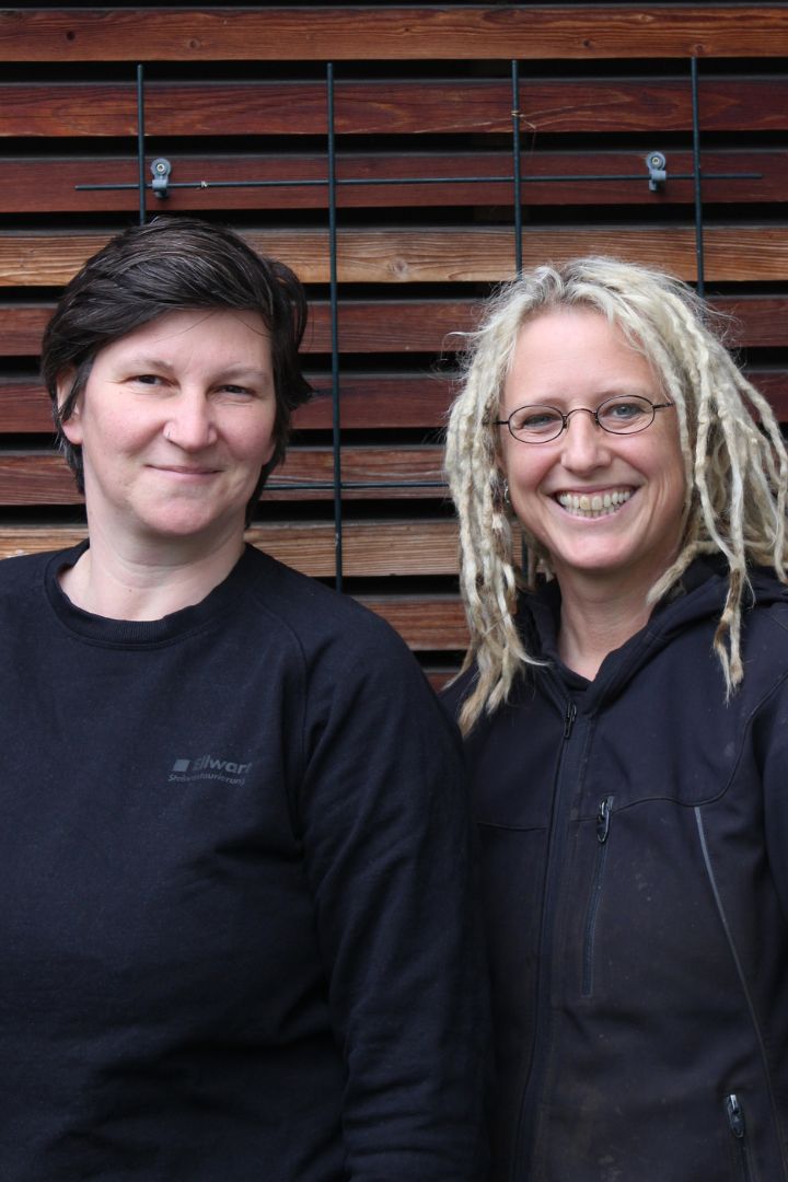 Tine Lippert und Gea Witt stehen vor einer Holzwand und lächeln herzlich in die Kamera. Sie bieten technische Restaurierung in Berlin und deutschlandweit an.