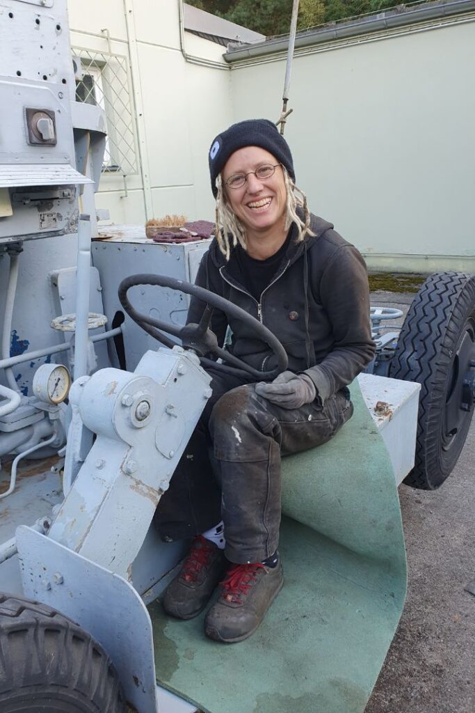 Restauratorin Gesa Witt bei der Restaurierung von technischem Kulturgut, lachend am Steuer eines historischen Fahrzeugs.